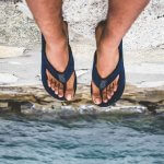 Legs and feet of a dark-skinned person in black flip flops seen dangling over a cliff with water below.